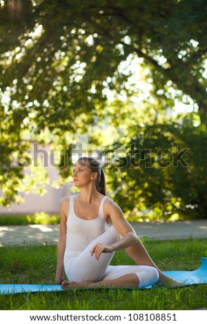 Beautiful young woman practices yoga in nature; sitting on carre-mat, twisted spine, holding her knee hand