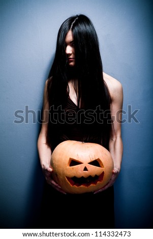 Gloomy young woman holding a large orange pumpkin carved for Halloween celebration. Dark background.