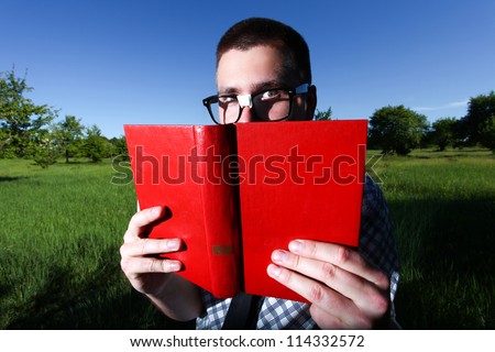 A handsome young man in glasses covered his face with a red book. Background of green field and blue sky