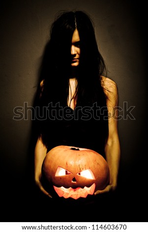 Gloomy woman holding a large glowing orange pumpkin carved for Halloween celebration. Dark background.