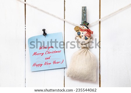 Christmas snowman clothespin hanging on clothesline or rope and holding sack and blue Christmas greeting note paper with push black pin on wood background