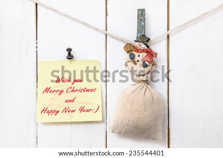 Christmas snowman clothespin hanging on clothesline or rope and holding sack and yellow Christmas greeting note paper with push black pin on wood background