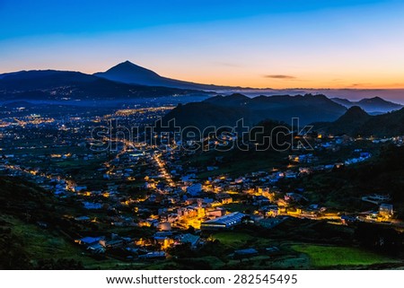 City or town with illumination after sunset or sundown at evening in mountains with blue sky and Teide volcano on background in Tenerife Canary island, Spain