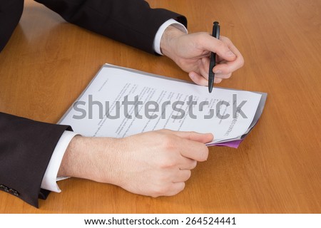 Businessman in dark suit and blue shirt sitting at office desk signing a contract with shallow focus on signature.