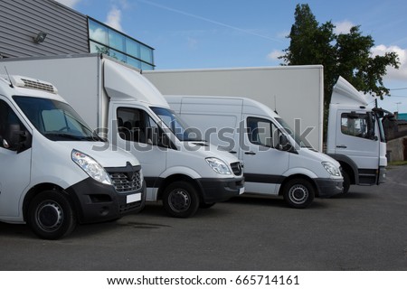Row of white delivery and service van, trucks and cars in front of a factory and warehouse distribution plant
