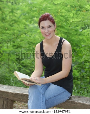 Young red haired woman reading a book on a wooden bench in a park
