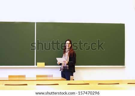 Teacher smiling, holding paper in empty classroom, blank blackboard in background