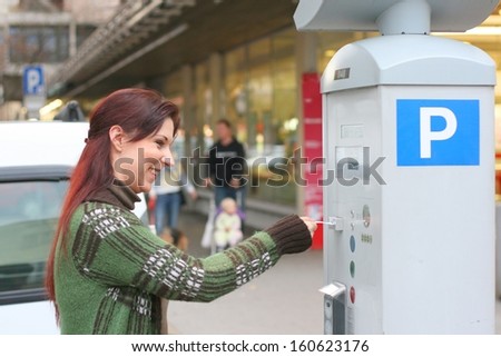 A young red haired woman paying for parking on machine in a street in a european city, people and supermarket in background