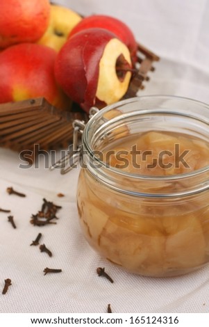 Home made apple compote in a jar (cooked apples in sugar water with spices) on white tablecloth, basket of apples and dried cloves