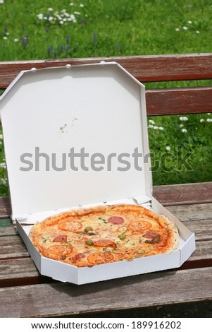 Pieces of pizza with fresh olives and tomato and salami in a white paper box, picnic on an old wooden bench in a park