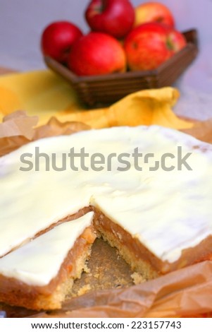 Apple pie with vanilla icing, one piece on cake server, apples in basket on yellow table cloth
