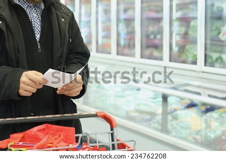 A man with shopping cart and list, grocery store refrigerator in background