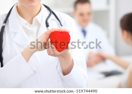 Female doctor with stethoscope holding heart.  Doctor and patient sitting in the background