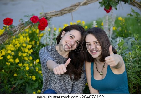 Two beautiful young women giving thumbs up sign and smiling on a hot summer day next to a flowers background