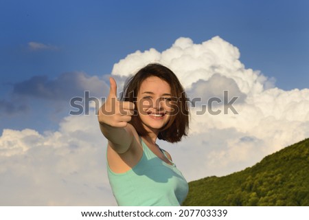 Beautiful young woman giving thumbs up sign in front of cloudy sky and smiling.