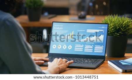 Female Analyst at Her Desk Works on a Laptop Showing Statistics, Graphs and Charts. She Works on the Wooden Table in Creative Office. Over the Shoulder Footage.