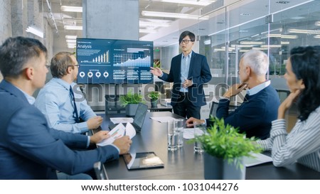 Asian Businessman Gives Report/ Presentation to His Business Colleagues, Pointing at the Results Showing Statistics, Pie Charts and Company\'s Growth On Wall TV Screen.