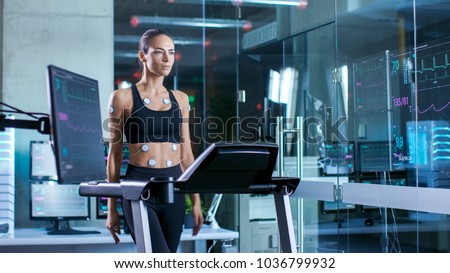 Beautiful Woman Athlete Wearing Sports Bra with Electrodes Attached to Her, Walks on a Treadmill in a Sports Science Laboratory. In the Background Laboratory with Monitors Showing EKG data.