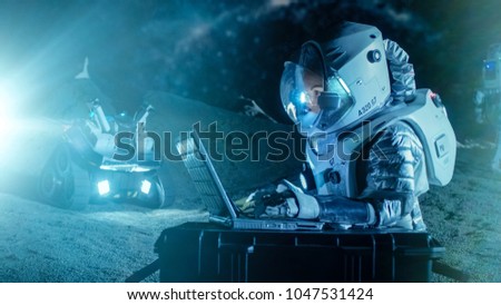 Female Astronaut Wearing Space Suit Works on a Laptop, Exploring Newly Discovered Planet, Communicating with the Earth. In the Background Her Crew Member and Space Habitat. Colonization Concept.