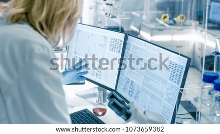 Senior Female Scientist Discusses Scientific Data with Her Laboratory Assistant. They\'re looking at Two Displays in a Modern Laboratory.