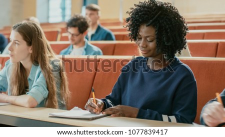 Beautiful Black Female Student Sitting Among Her Fellow Students in the Classroom, She\'s Writing in the Notebook and Listens to a Lecture.