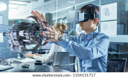 Computer Science Engineer wearing Virtual Reality Headset Works with 3D Model Hologram Visualization, Makes Gestures. In the Background Engineering Bureau with Busy Coworkers.