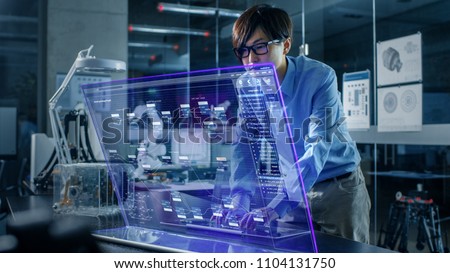 Asian Neural Microchip Design Engineer Uses Modern Computer With Transparent Holographic Display. Monitor Shows Advanced Technology Interface . Shot in Modern Glass and Concrete Office.