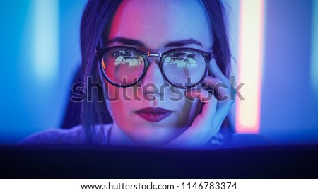 Portrait of the Beautiful Young Girl Sitting Before Computer Screen, Browsing in Internet, Playing Online Games, Streaming. Cute Girls Wearing Glasses in the Cool Retro Neon Lit Room.