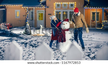 Happy Young Couple Making Snowman in the Backyard of their Idyllic House while it is Snowing. They Wrap up Snowman with Scarf. Family Spending Time Together on Christmas Eve.