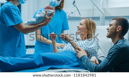 In the Hospital Midwife Gives Newborn Baby to a Mother to Hold, Supportive Father Sitting Near Wife. Happy Family in the Modern Delivery Ward.