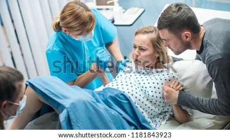 In the Hospital, Woman in Labor Gives Birth, Obstetricians and Doctors Assist, Her Husband Supports Her by Holding Hand. Modern Maternity/ Delivery Ward with Professional Midwives.