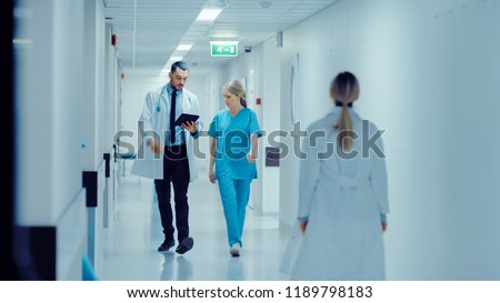Female Surgeon and Doctor Walk Through Hospital Hallway, They Consult Digital Tablet Computer while Talking about Patient\'s Health. Modern Bright Hospital with Professional Staff.