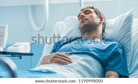 In the Hospital Sick Male Patient Lies on a Bed Sleeping, Heart Rate Monitor On His Finger. Bright and Modern Medical Ward.