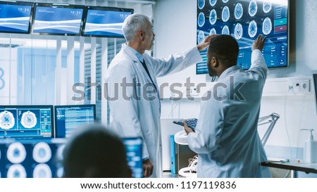 Team of Scientists Work in the Brain Research Laboratory, Discussing Brain Scans Show on Wall TV Monitor. Neurologists / Neuroscientists Surrounded by Monitors Showing CT, MRI Scans.