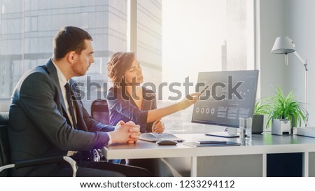 Female Top Manager and Male Businessman Sitting at the Desk Having Discussion and Working on a Desktop Computer, Solving Problems. Successful People in Modern Stylish Office with Big City Busines View