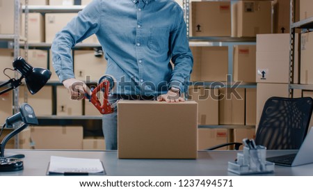 Professional Warehouse Worker Checks and Seales Cardboard Box Ready for Shipment. In the Background Person Working in the Rows of Shelves with Cardboard Boxes with Ready Orders.