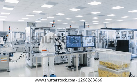 Shot of Sterile High Precision Manufacturing Laboratory where Scientists in Protective Coverall\'s Use Computers and Microscopes, doing Pharmaceutics, Biotechnology and Semiconductor Research.