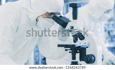 In Laboratory Scientist in Protective Clothes Doing Research, Using Microscope and Writing Down Data. Workers Working on a Modern Manufactory Producing Semiconductors and Pharmaceutical Items.