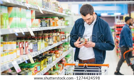 At the Supermarket: Handsome Man Uses Smartphone and Looks at Nutritional Value of the Canned Goods. He\'s Standing with Shopping Cart in Canned Goods Section.
