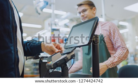 At the Supermarket: Checkout Counter Customer Pays with Smartphone for His Food Items. Big Shopping Mall with Friendly Cashier, Small Lines and Modern Wireless NFC Paying Terminal System.