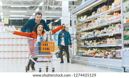 At the Supermarket: Man Pushes Shopping Cart with Woman Sitting in it, Happy Couple Has Fun Racing in a Trolley through the Fresh Produce Section of the Store.