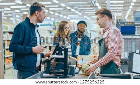 At the Supermarket: Checkout Counter Customer Pays with Smartphone for His Items. Big Shopping Mall with Friendly Cashier, Small Lines and Modern Wireless Paying Terminal System.