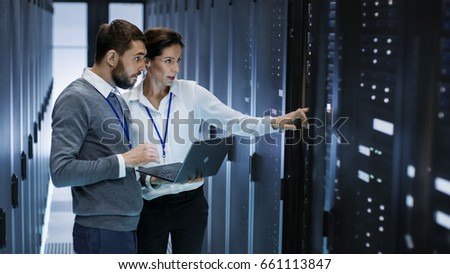 Male IT Specialist Holds Laptop and Discusses Work with Female Server Technician. They\'re Standing in Data Center, Rack Server Cabinet is Open.