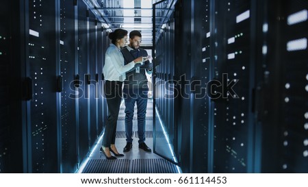 Female IT Technician and Male Server Engineer Talk and Discuss Settings of a Working Data Center. Man Holds Laptop.