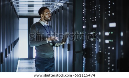 IT Technician Works on Laptop next to a Server Cabinet in Big Data Center. He Runs Diagnostics and Maintenance, Sets System Up.