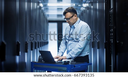 In Data Center IT Engineer Works on Crash Cart Laptop. We See Rows of Server Racks and One Cabinet is Open.