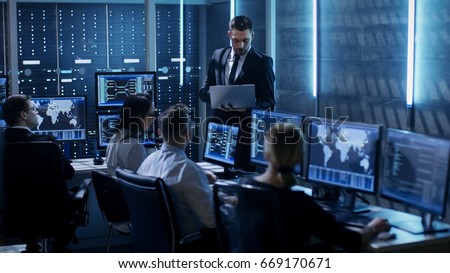 Professional IT Engineers Working in System Control Center Full of Monitors and Servers. Supervisor Holds Laptop and Holds a Briefing. Possibly Government Agency Conducts Investigation.