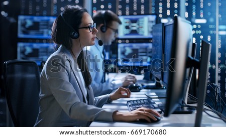 Female working in a Technical Support Team Gives Instructions with the Help of the Headsets. In the Background People Working and Monitors Show Various Information.