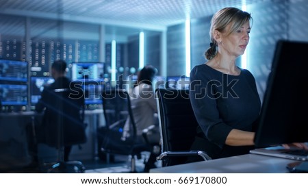 Female IT Engineer Works on Her Desktop Computer in Government Surveillance Agency. In the Background People at Their Workstations with Multiple Screens Showing Graphics.