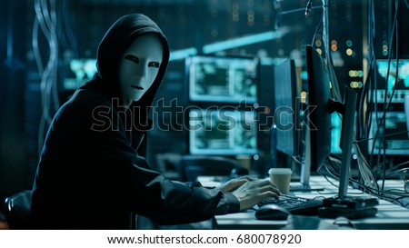 Masked Hacker is Using Computer for Organizing Massive Data Breach Attack on Corporate Servers. They\'re in Underground Secret Location Surrounded by Displays and Cables.